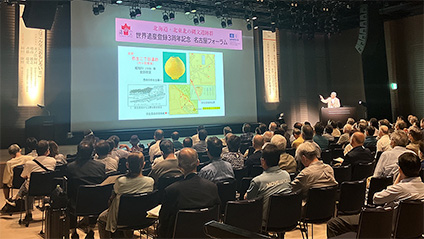 프로젝터 화면에 영상이 비춰지고 연사가 연설하고 있다. 의자에 앉아 있는 사람들이 프로젝터 화면을 보고 있다.