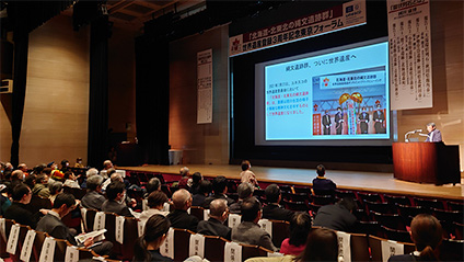 东京论坛的现场画面投射在屏幕上，演讲者正在发言